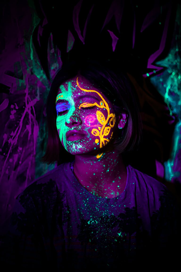 A woman with neon face paint glows in dark surroundings, closed eyes enhancing artistic effect.